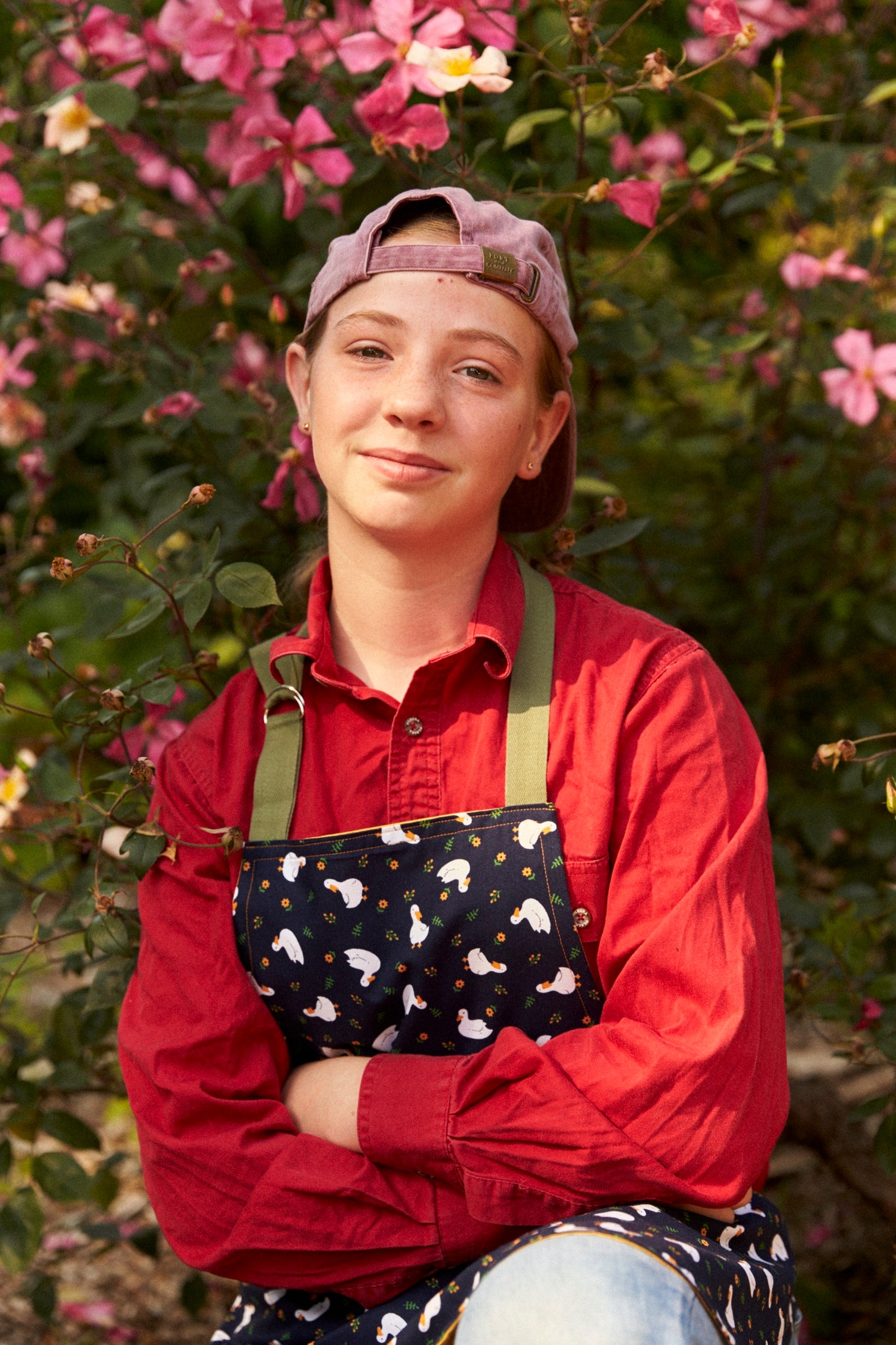 Person wearing a red shirt and apron with a floral pattern, standing in front of pink flowers.