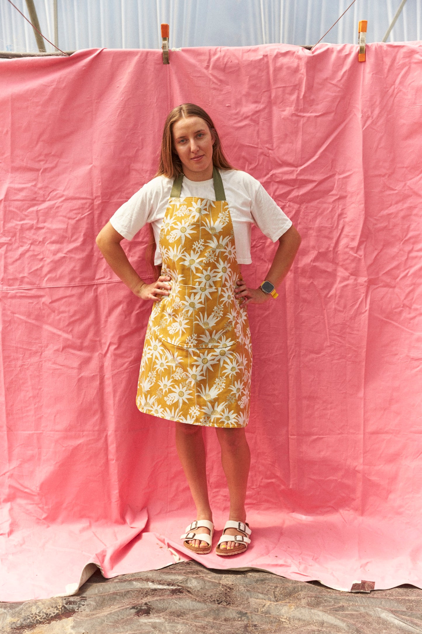 Person wearing a yellow floral apron over a white shirt against a pink backdrop