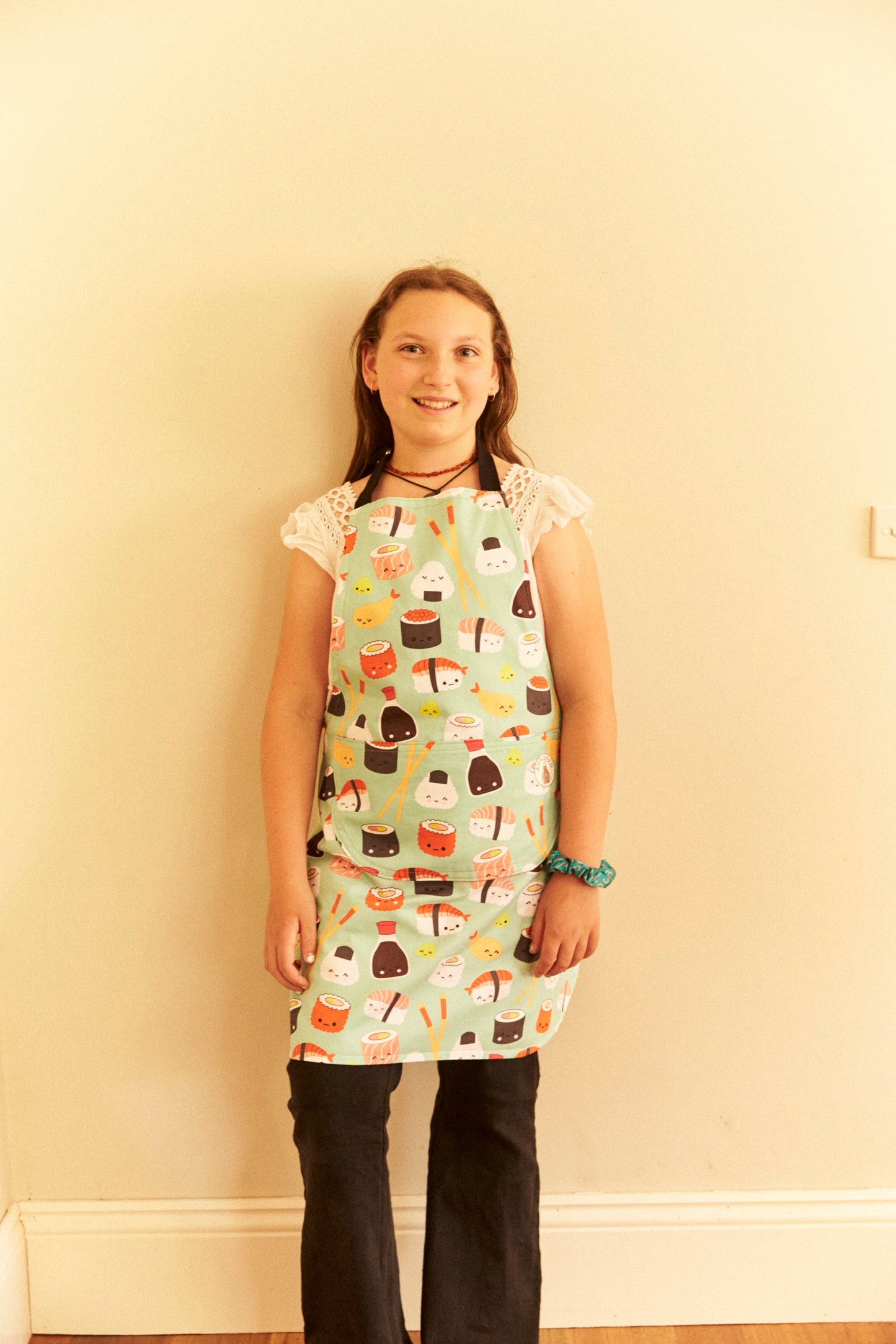 Young girl wearing a colorful apron with food illustrations against a plain wall.
