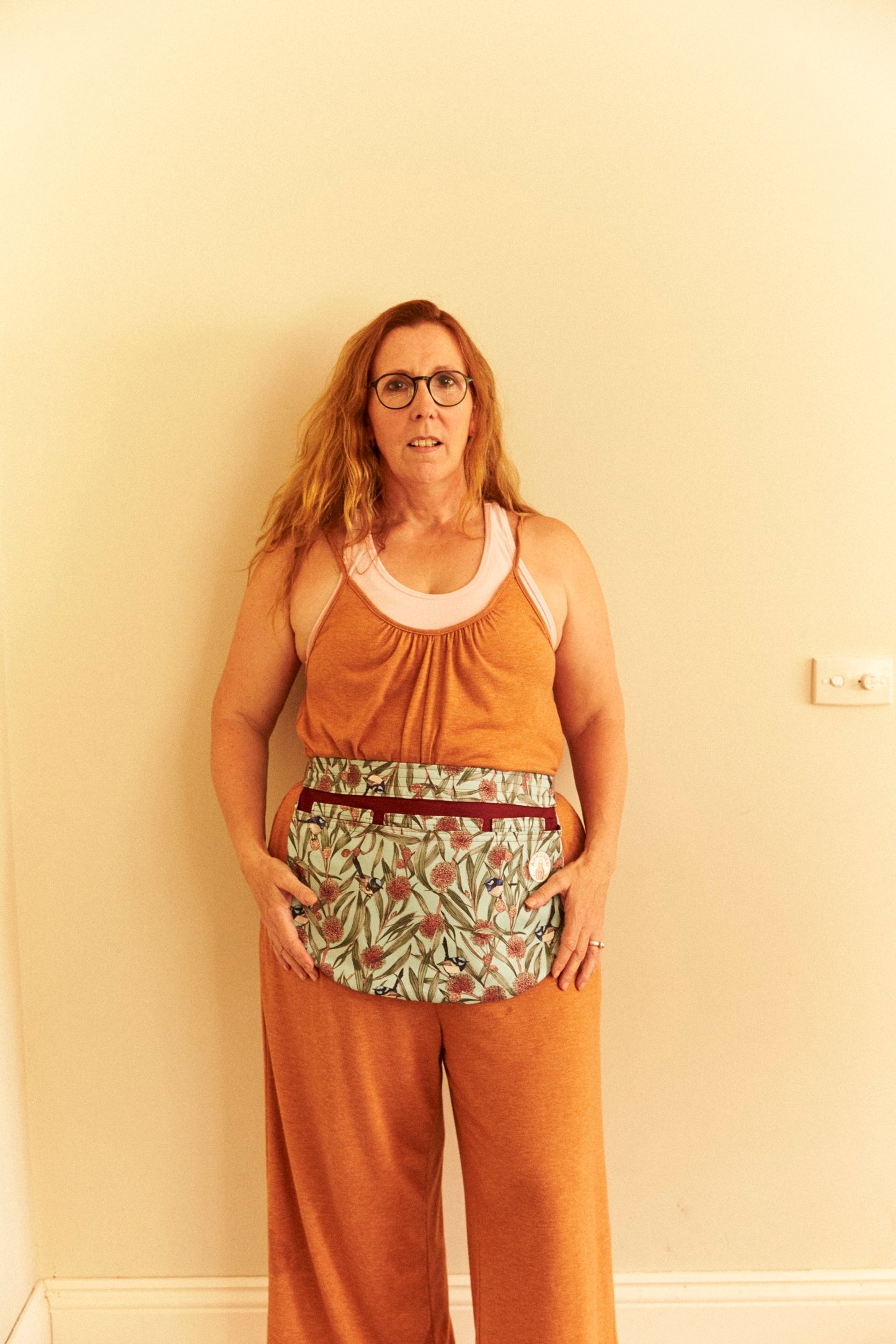 Woman wearing a rust-colored sleeveless top and pants with a floral pattern against a beige wall.