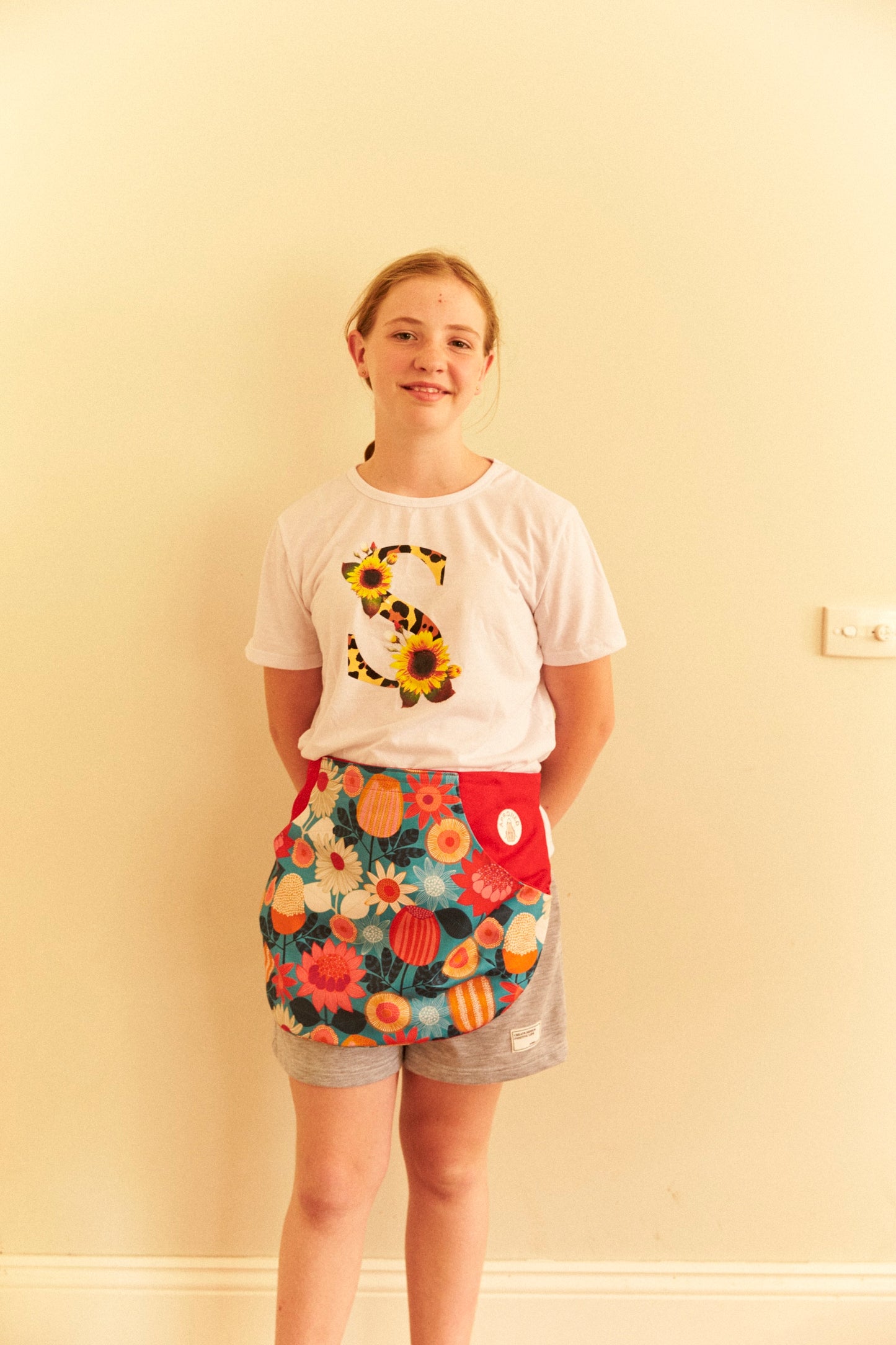 Person wearing a white t-shirt with a sunflower design and colorful floral shorts against a plain wall.