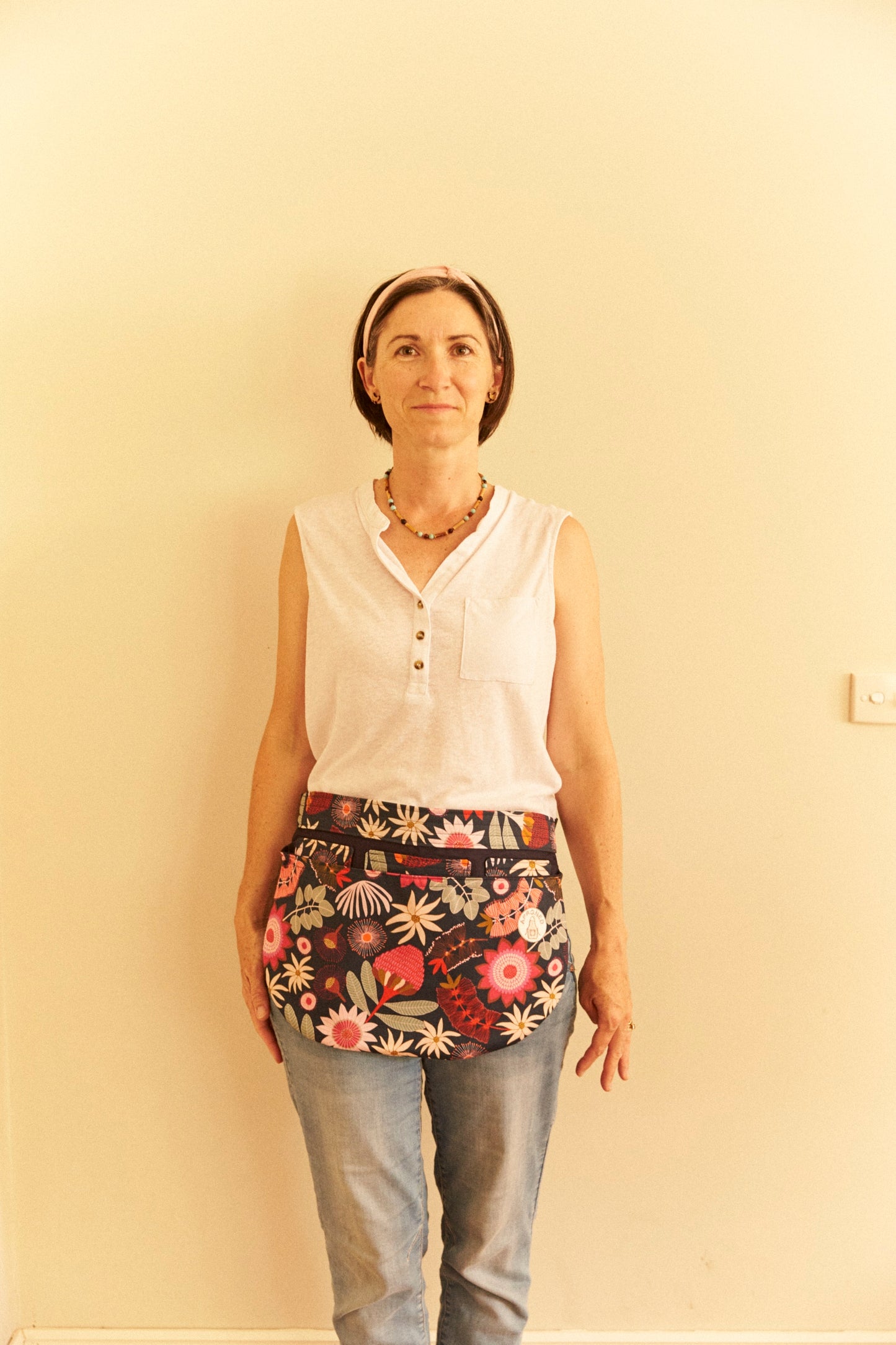 Woman wearing a white sleeveless top and floral apron against a plain wall.