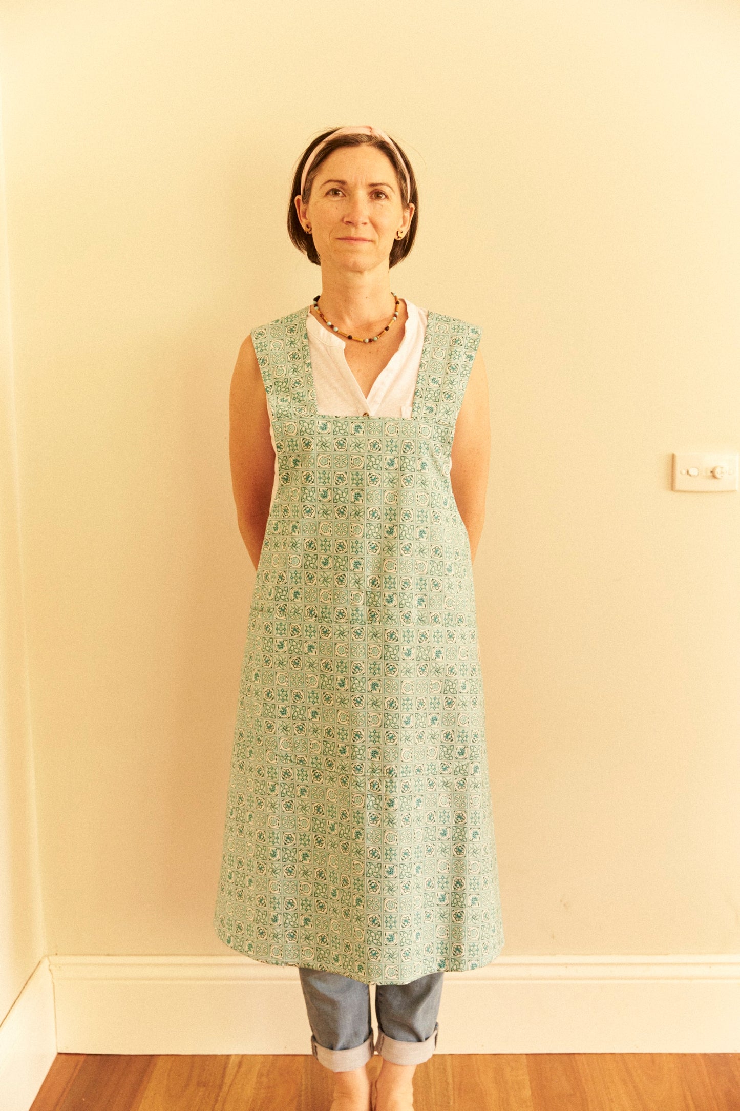 Woman wearing a green floral apron over a white shirt and blue jeans against a beige wall.