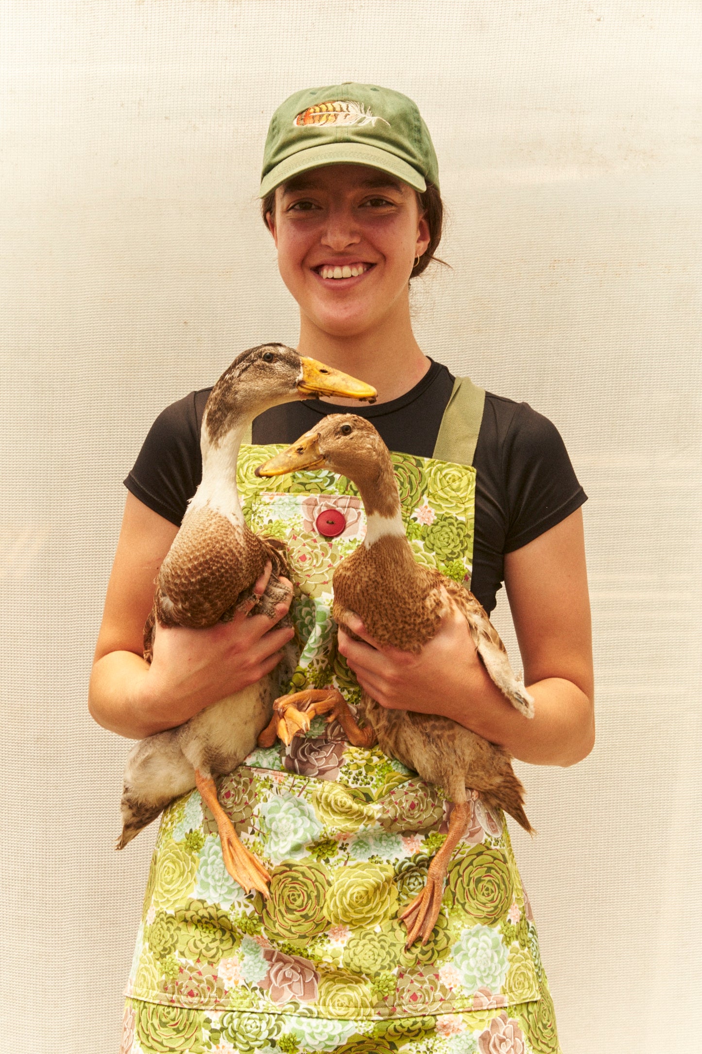 Person holding two ducks wearing a green cap and apron against a plain background