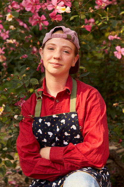 Person wearing a red shirt and apron with a floral pattern, standing in front of pink flowers.