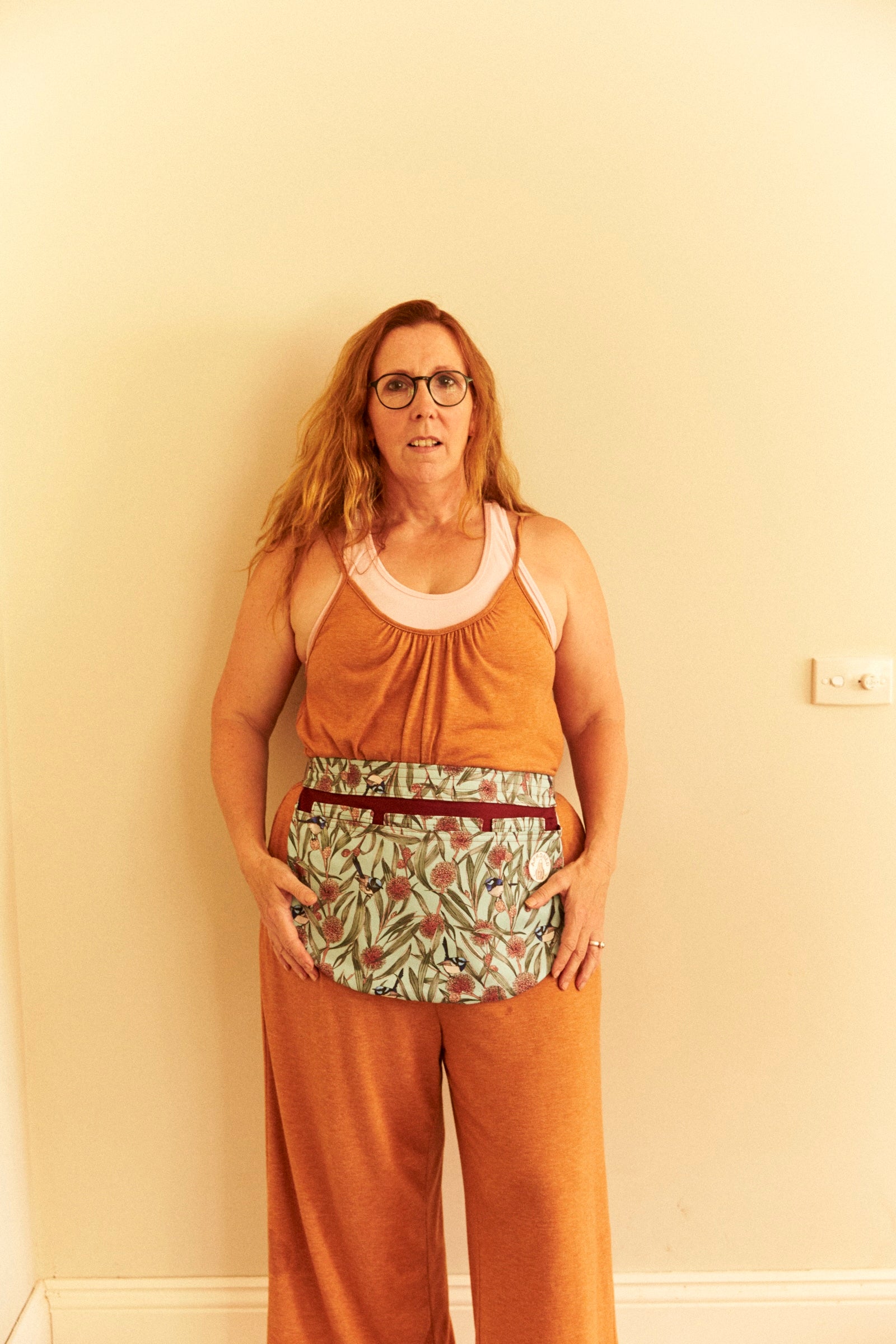Woman wearing a rust-colored sleeveless top and pants with a floral pattern against a beige wall.