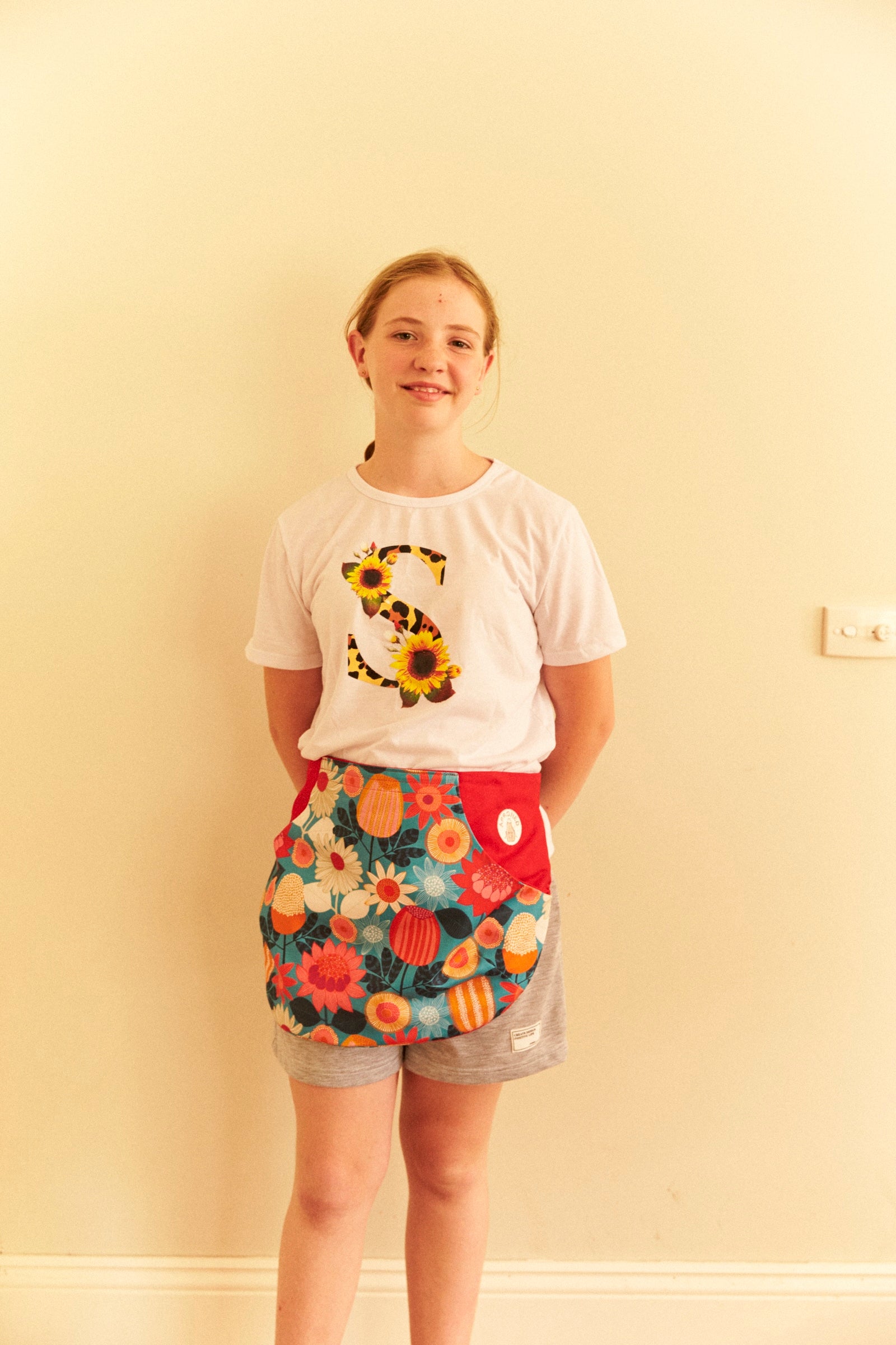 Person wearing a white t-shirt with a sunflower design and colorful floral shorts against a plain wall.