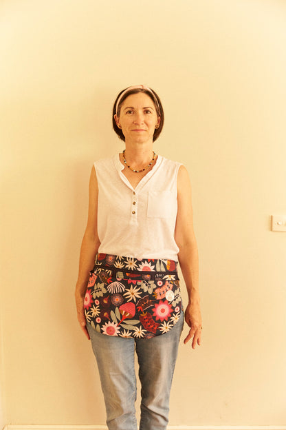 Woman wearing a white sleeveless top and floral apron against a plain wall.