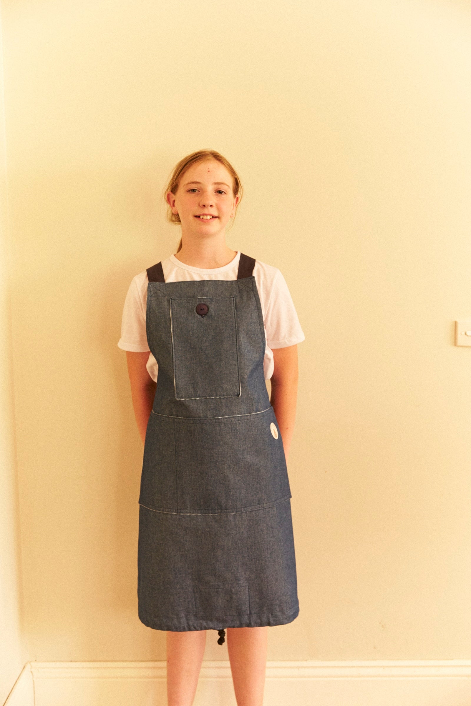 Person wearing a denim pinafore dress against a plain wall.