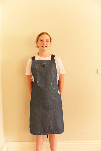 Person wearing a denim pinafore dress against a plain wall.