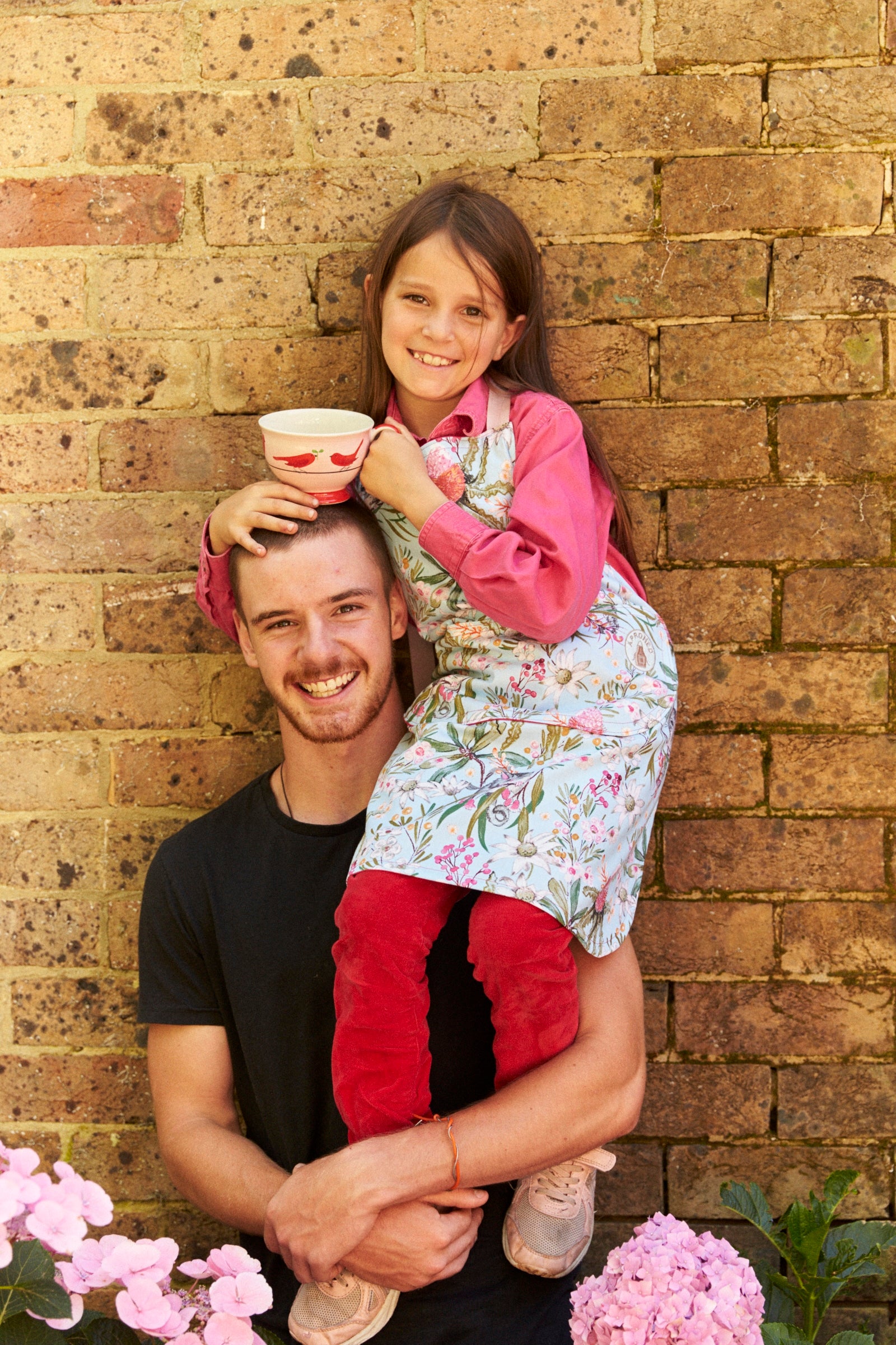 Man holding a young girl on his shoulders against a brick wall with pink flowers in the foreground.