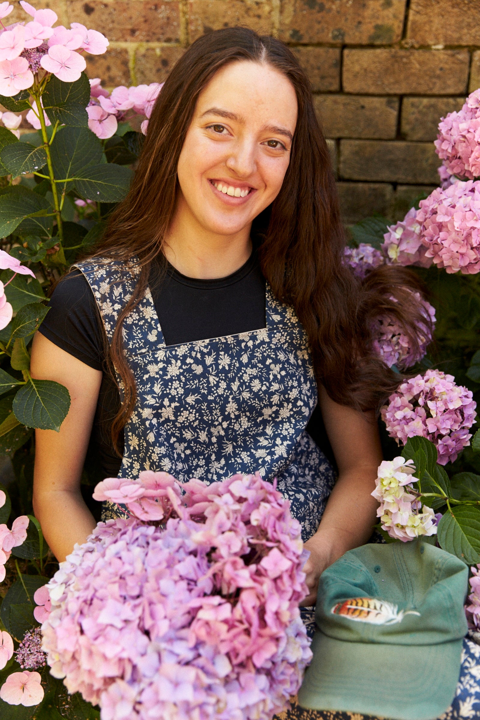 Woman in a floral apron sitting among pink flowers with a brick wall background