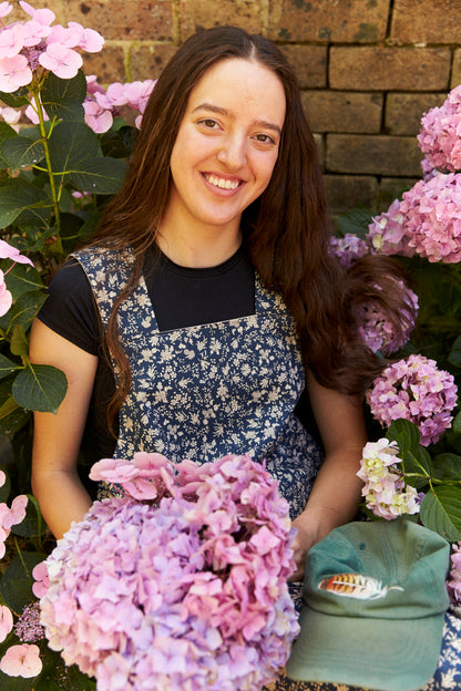 Woman in a floral apron sitting among pink flowers with a brick wall background