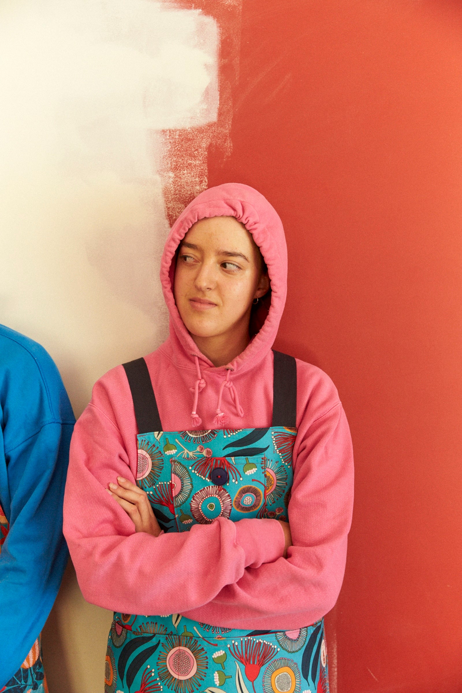 Person wearing a pink hoodie and colorful apron standing against a red wall.