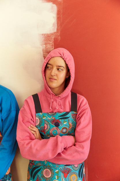 Person wearing a pink hoodie and colorful apron standing against a red wall.