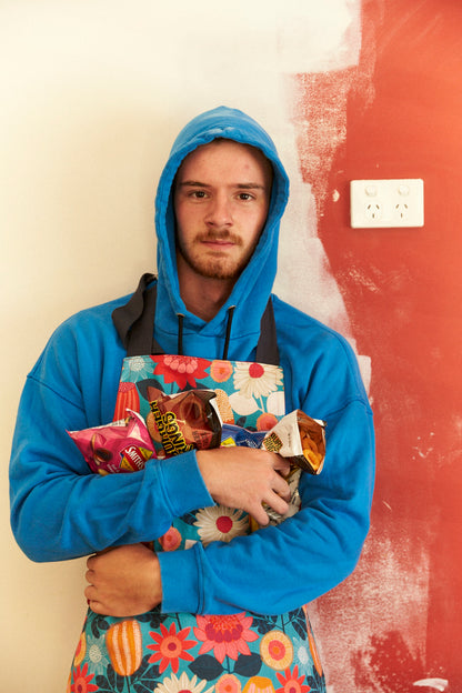 Person wearing a blue hoodie holding colorful bags against a red wall.