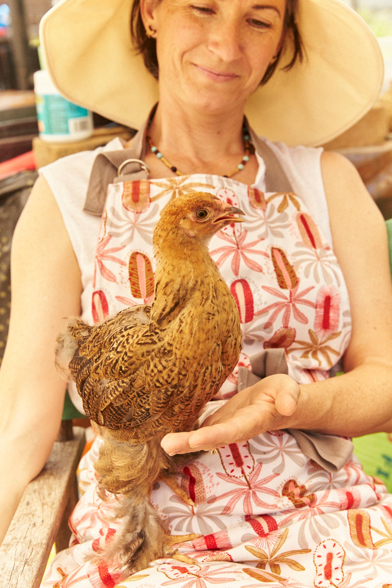 Woman holding a chicken wearing a floral apron and wide-brimmed hat.