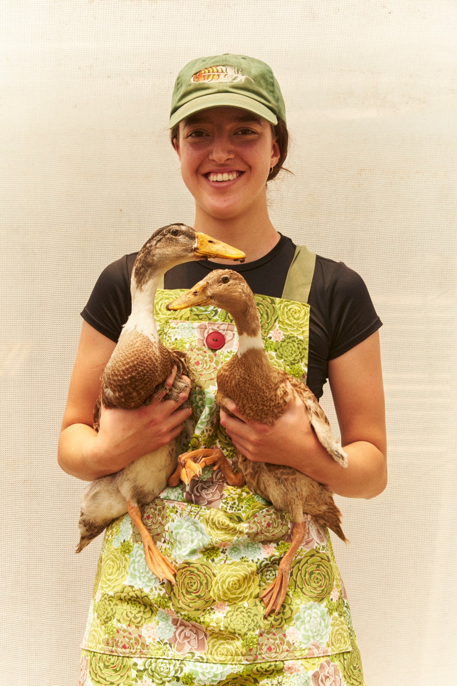 Person holding two ducks wearing a green cap and apron against a plain background