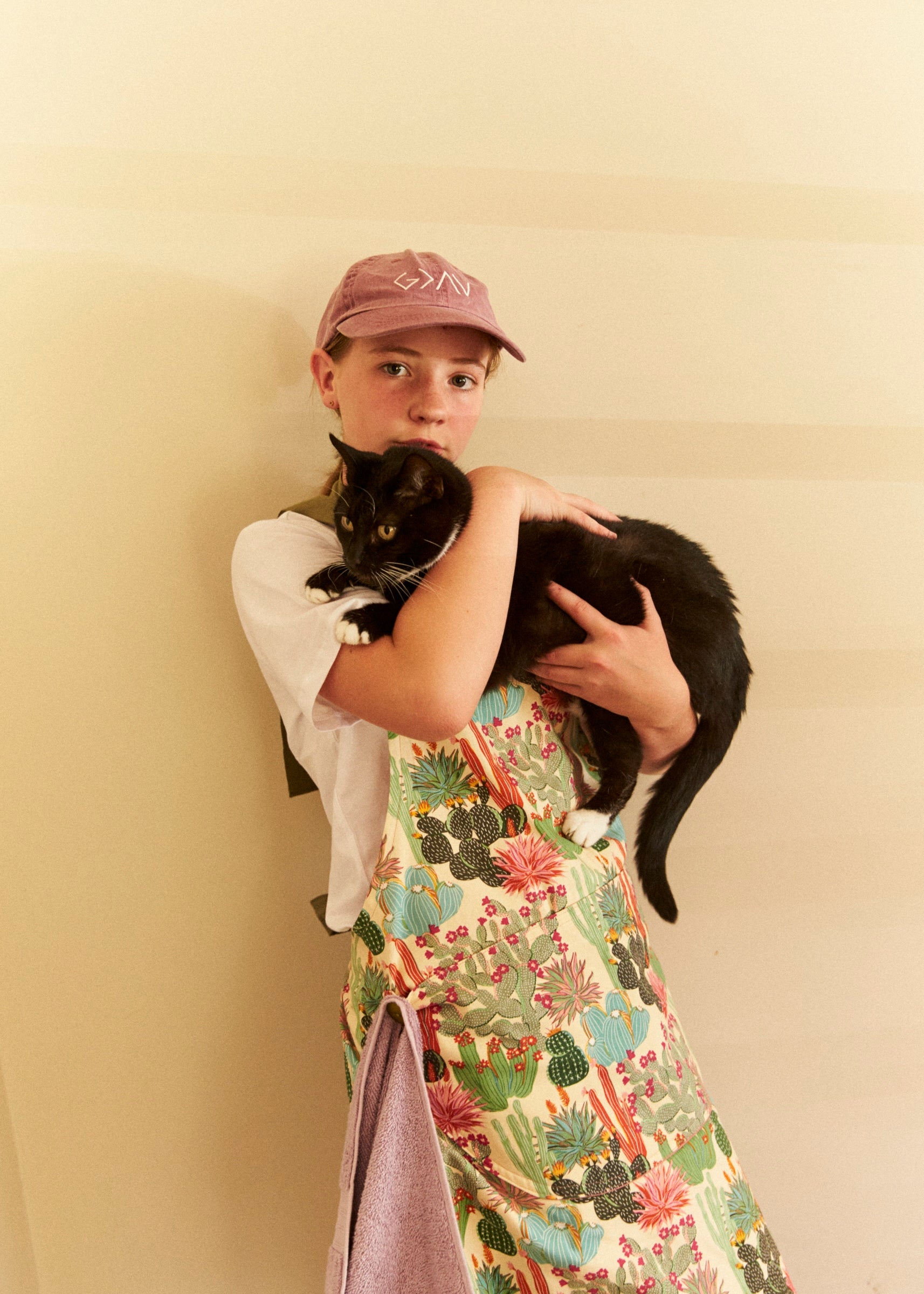 Person wearing a colorful apron and cap holding a black cat against a beige wall.